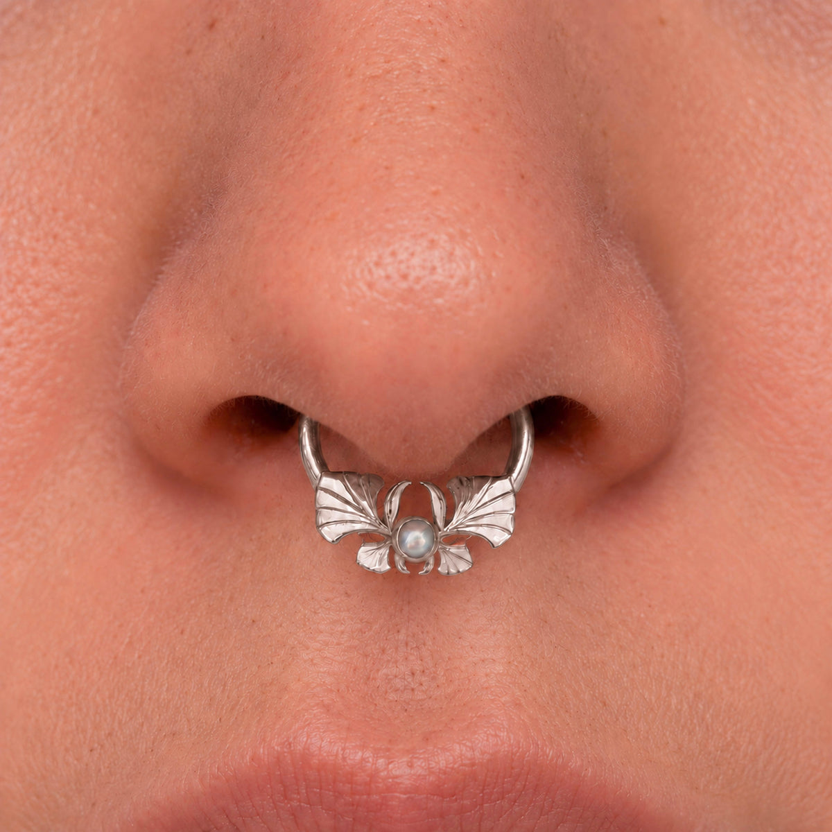 Silver septum piercing ring with a pearl and ornate wing design on a person's nose