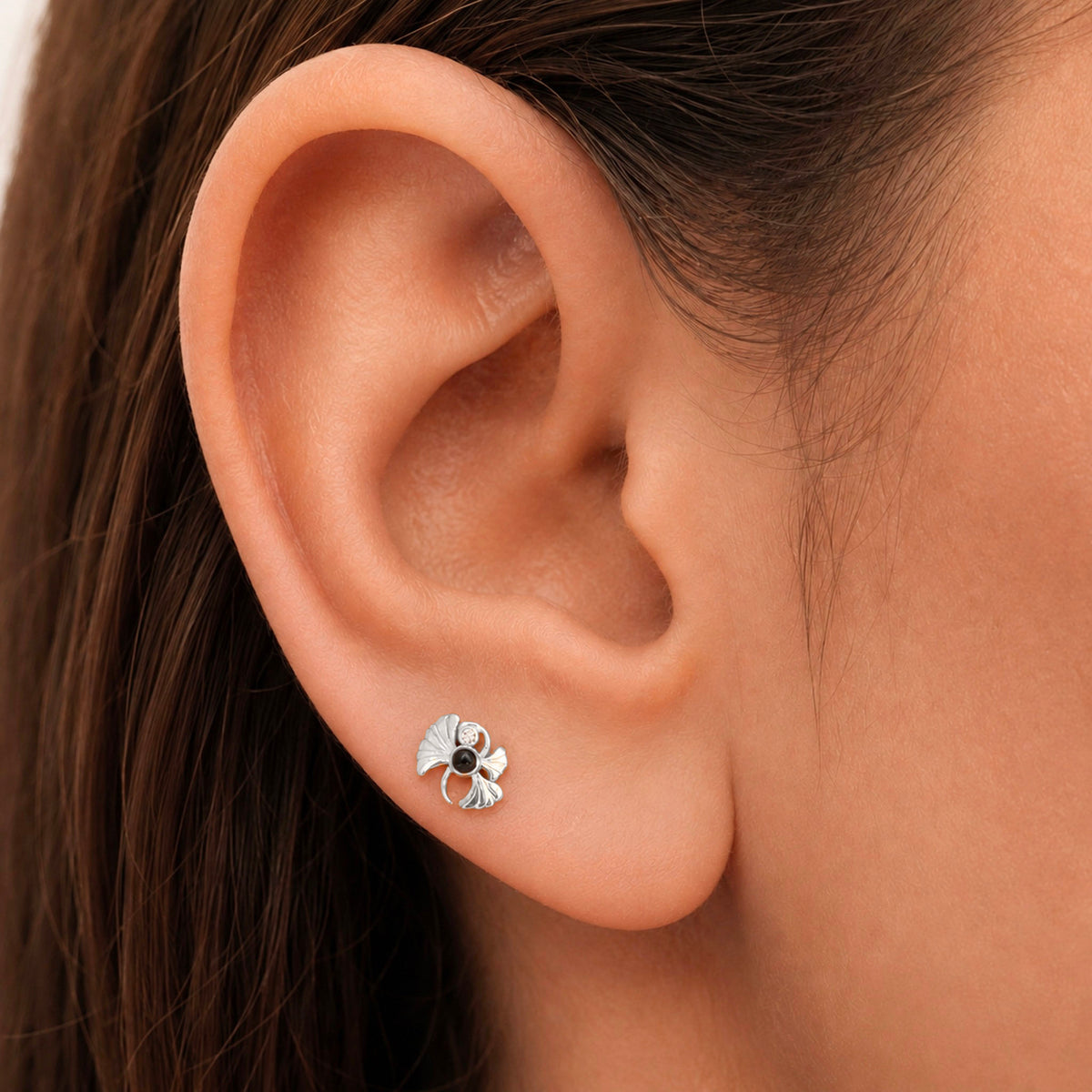 Close-up of a silver ginkgo leaf stud earring with a black gemstone in a human ear.