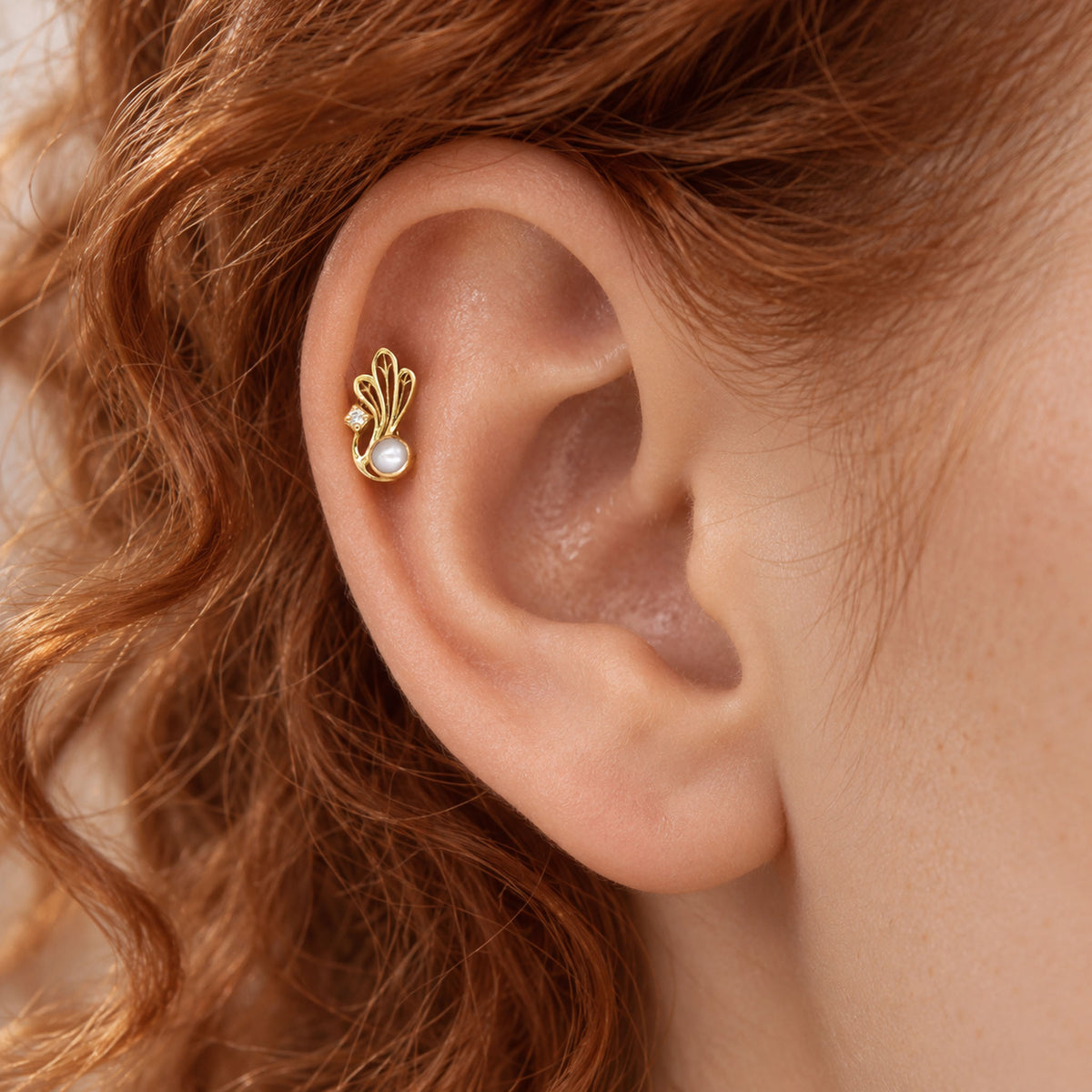Close-up of a gold helix piercing with a pearl and diamond leaf design on a redhaired woman's ear