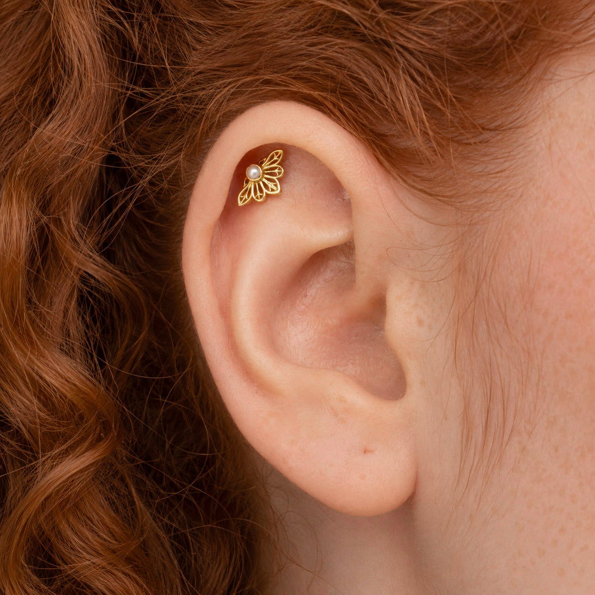 Close-up of an 18k gold flower-shaped helix piercing with a small pearl on a human ear.