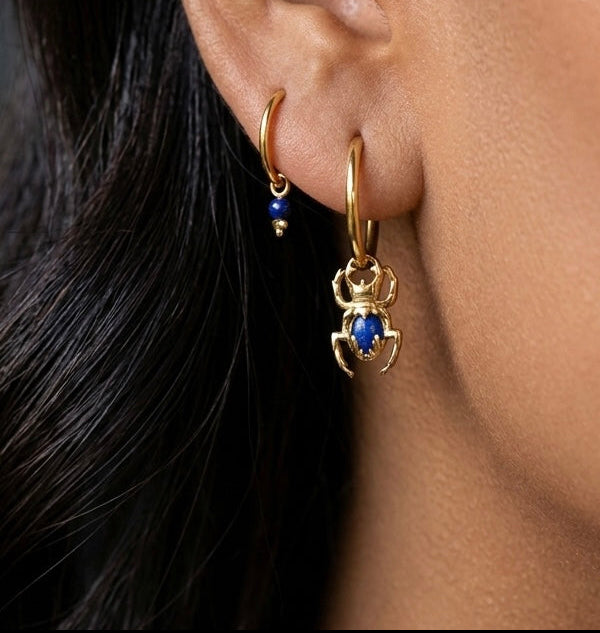 Close-up of ear with 18k gold hoop earrings, one featuring a blue gemstone scarab charm.