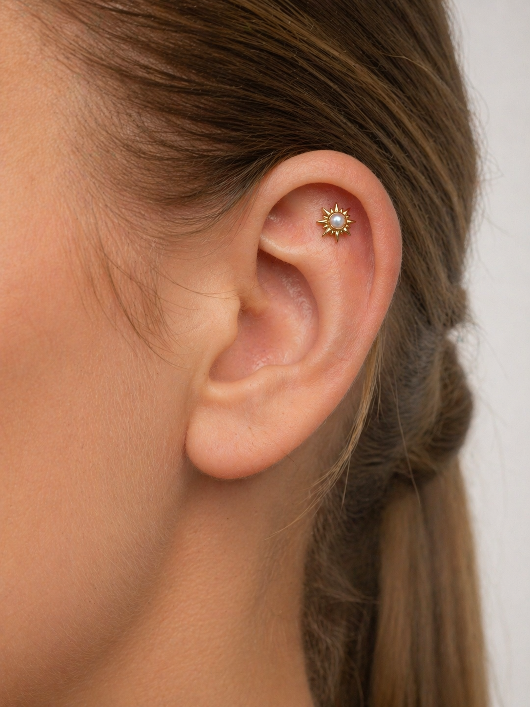 Close-up of ear with 18k gold sun-shaped cartilage piercing with pearl center